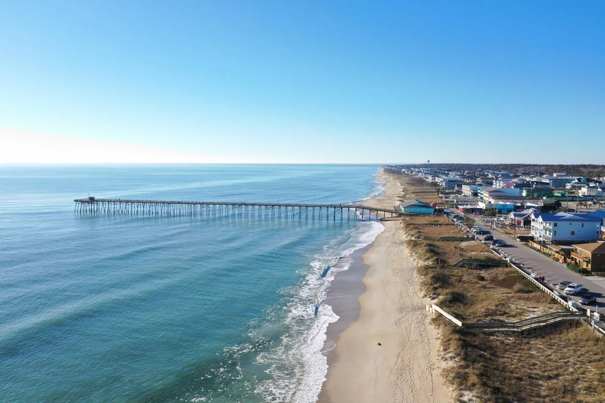 The iconic Kure Beach Pier stretches out over the ocean, surrounded by sandy shores and charming coastal homes. Visitors stroll the pier, fish, and enjoy the soothing sounds of the waves, encapsulating the diverse outdoor activities available to guests of all ages.