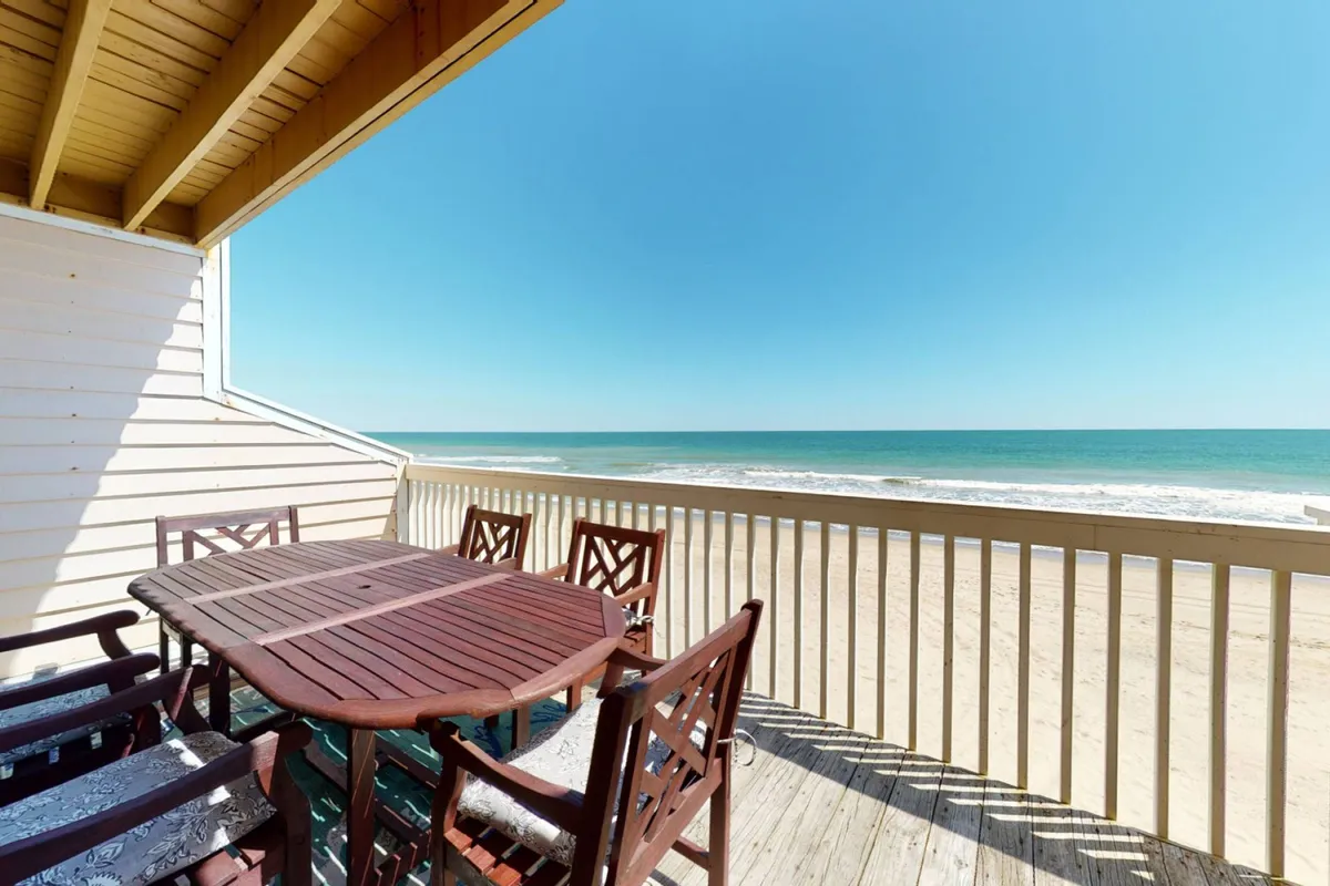 A wide, sunlit deck with an outdoor dining table and chairs overlooks the tranquil ocean and sandy beach. The space exudes relaxation, with room for group gatherings, sunrise coffee moments, and evening barbecues, all set against a stunning ocean backdrop.