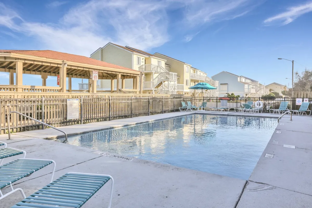 An exterior view of the resort's outdoor swimming pool area, featuring lounge chairs, a covered seating space, and adjacent buildings. The scene feels vibrant and inviting, with spaces for both sunbathing and socializing. The layout suggests ample room for families and friends to gather and enjoy the outdoors.