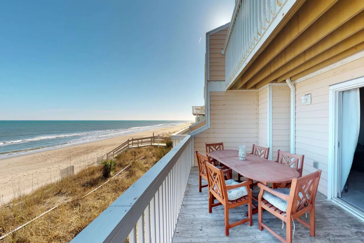 An exterior view from the oceanfront deck, featuring a dining table and chairs overlooking the ocean through a glass railing. The scene shows the transition from indoor to outdoor living, inviting al fresco meals or sunset gatherings with unobstructed water views.
