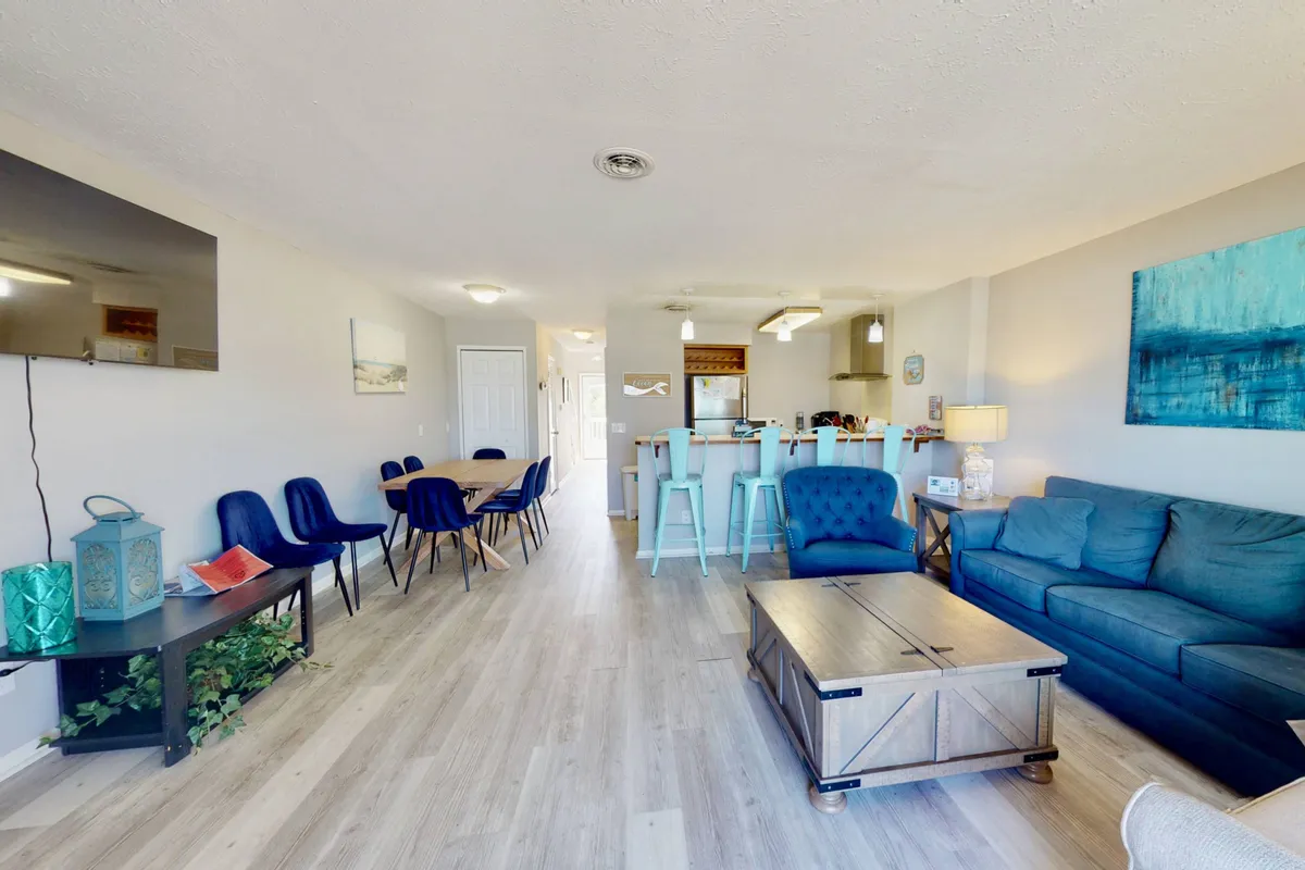 A wide-angle view of a bright, open-concept living space, featuring a blue sofa, wooden coffee table, kitchen bar area with stools, and adjacent dining area with chairs. Large windows flood the room with natural light, and the seamless flow between the kitchen, dining, and living areas is immediately apparent.