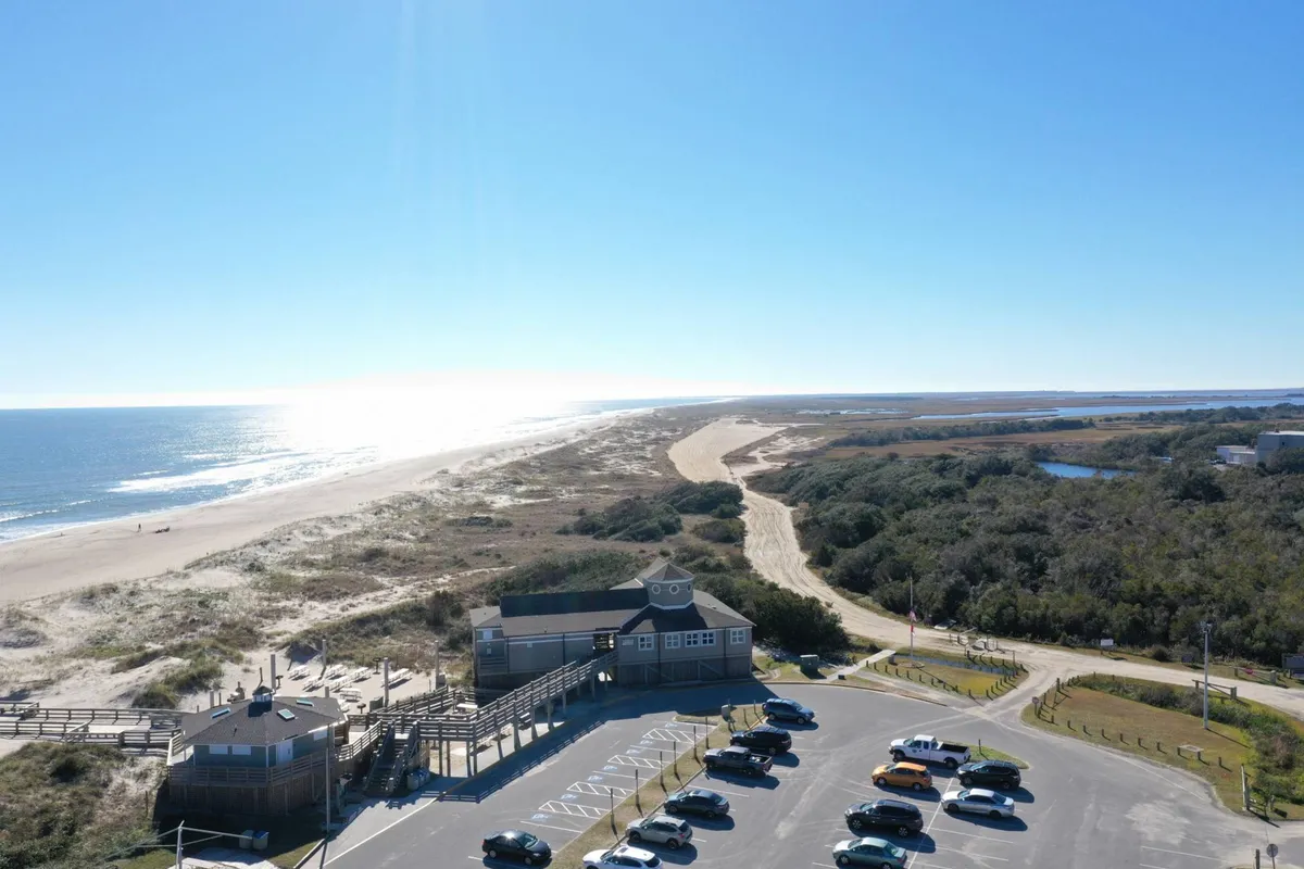 A broad view of the ocean, sand dunes, parking lot, and the beach access building. The scene captures the feeling of arrival at a vibrant, amenity-rich destination, suggesting community events, outdoor activities, and the convenience of resort-style access to the beach.
