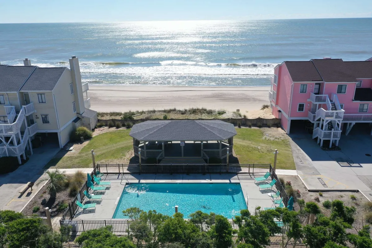 The hero image showcases an inviting beachfront swimming pool surrounded by lush greenery, a charming gazebo perfect for gathering, and vibrant condos overlooking the ocean. The scene exudes a sense of relaxation and community, with the sandy beach and waves just steps away, ideal for both peaceful moments and lively family fun.