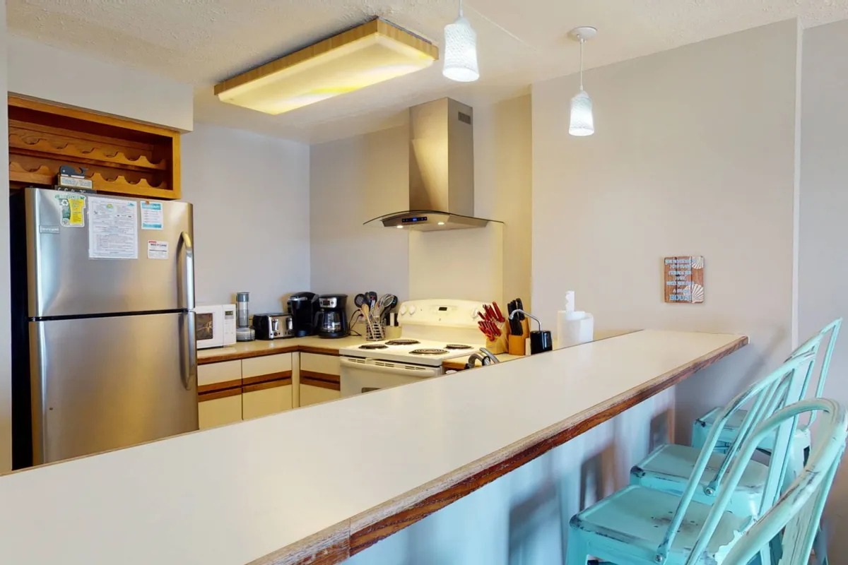 A welcoming kitchen scene with a central island, stainless steel appliances, and colorful bar stools, all bathed in warm light. The kitchen is open to the living space, inviting group cooking, conversation, and shared meals.