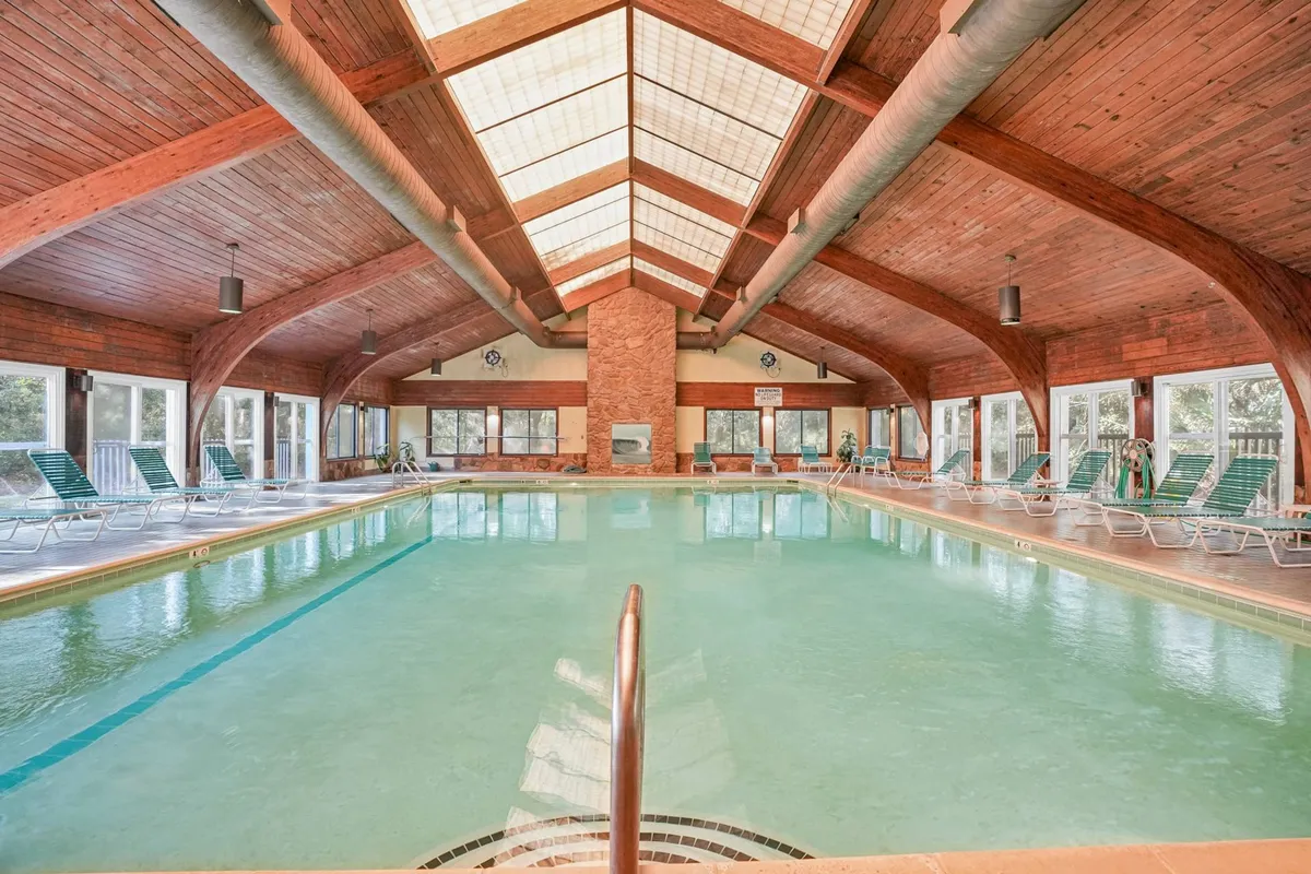 A bright and inviting indoor swimming pool area with wooden beams, large windows letting in sunshine, and comfortable lounge chairs. The open design suggests a place where families can enjoy swimming and relaxation regardless of the weather.