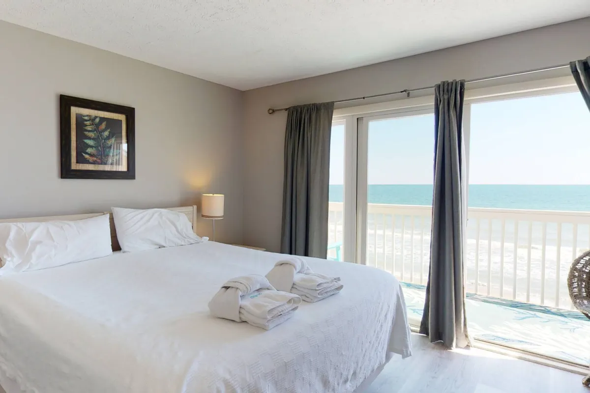 A tranquil bedroom with a large bed positioned near a window that offers an expansive ocean view. A lamp and tasteful framed artwork add a touch of coziness, while the serene color palette and natural light create a restful ambiance. The proximity to the ocean and access to a balcony are evident, suggesting a private outdoor retreat.