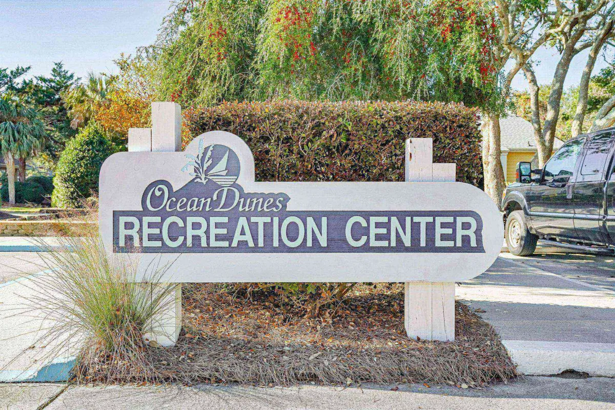 The Ocean Dunes Recreation Center sign is set amidst lush greenery, signaling the availability of nearby community amenities and gathering spaces. Vehicles in the background hint at easy accessibility for all family members.