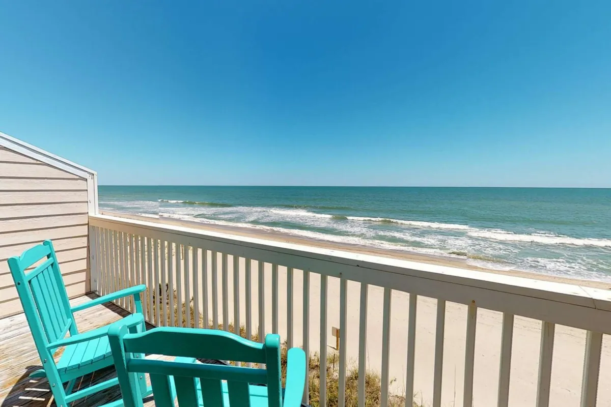 A serene exterior view featuring inviting turquoise chairs on a deck overlooking a pristine white sandy beach and expansive ocean. The setting suggests relaxation, sunrise views, and proximity to the beach—the heart of the vacation experience.