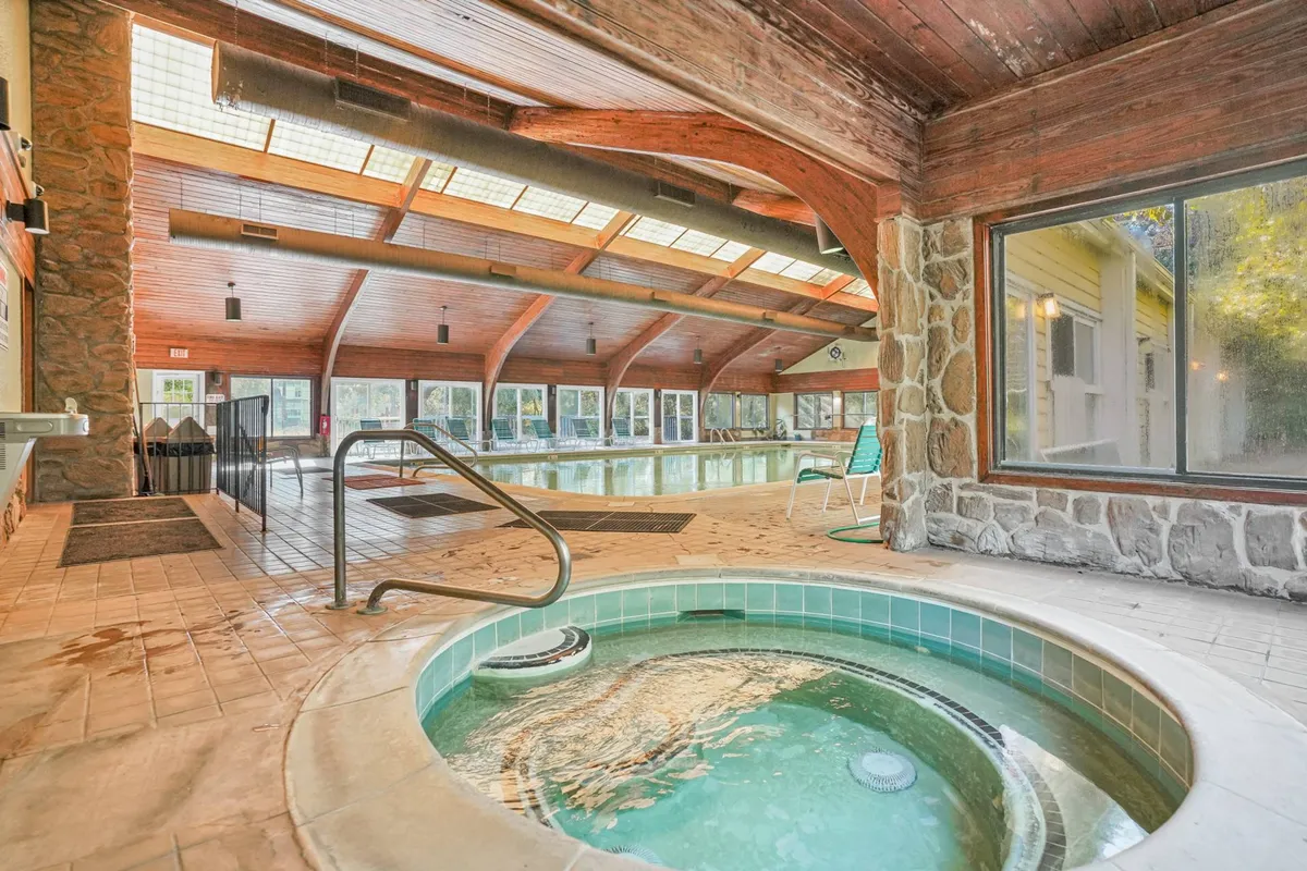 This inviting interior scene highlights an indoor pool and hot tub area, framed by large windows that let in natural light. Wooden beams and stone walls add warmth and a touch of rustic charm. Guests are seen swimming, relaxing in the hot tub, and socializing, capturing both active and leisurely recreation.