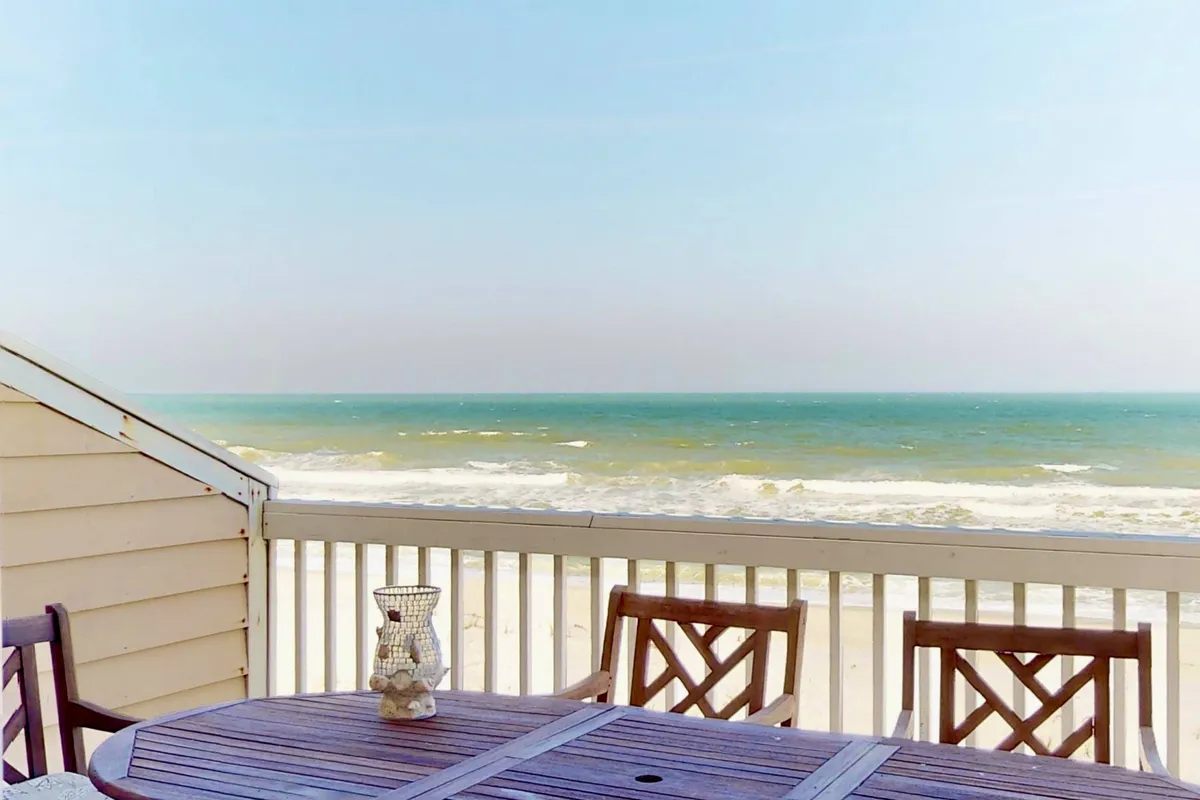 This image showcases an exterior deck overlooking the ocean, featuring a wooden dining table and beach chairs. The scene suggests a place where guests can enjoy outdoor meals, cocktails, or simply relax with friends while soaking up tranquil sea views. The presence of the ocean in the background highlights the close proximity to the beach and the laid-back, social atmosphere of the resort townhome.